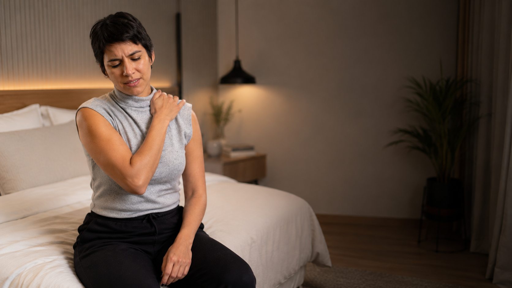 Mulher sentada na cama segurando o ombro com dor